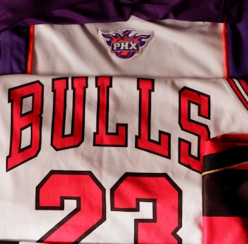 The image features a collection of sports jerseys, including a purple jersey with an emblem, a white jersey with red lettering spelling 'BULLS' and the number 23, and a black and white striped jersey with the Juventus emblem.